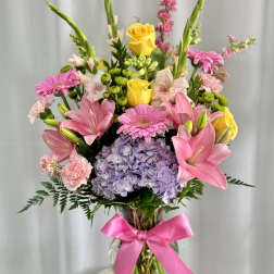 Mixed bouquet of pink lilies, yellow roses, and lavender hydrangea in a glass vase with a pink ribbon