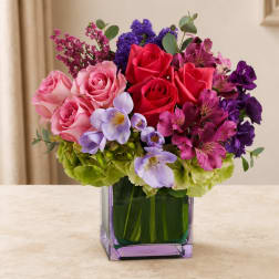 Mixed bouquet of pink, red, purple, and lavender flowers in a square glass vase