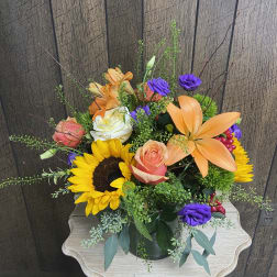Mixed bouquet with sunflowers, orange lilies, roses, and purple blooms in a glass vase