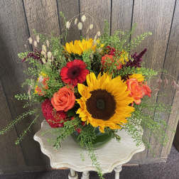 Low arrangement of sunflowers, orange roses, and red blooms in a green bowl on a small table