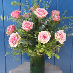 Tall arrangement of pink roses with airy filler in a clear glass vase on a small wooden table