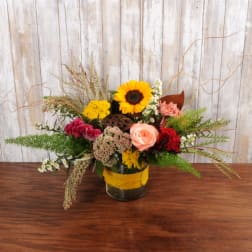 Low rustic arrangement with sunflower, peach rose and red blooms in a glass vase with yellow wrap