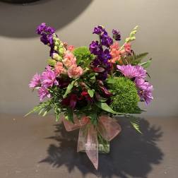Mixed bouquet of purple, pink, and green flowers in a glass vase with a pink ribbon