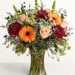 Mixed bouquet of orange gerbera daisies, peach roses, and burgundy blooms in a glass vase