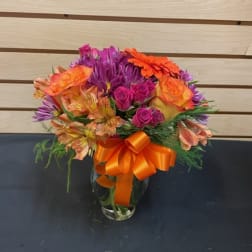 Colorful bouquet of roses, alstroemeria, and purple daisies in a glass vase with an orange ribbon