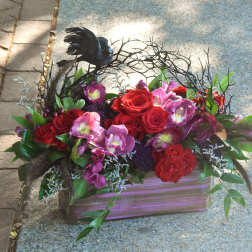 Low rectangular arrangement of red roses and purple flowers with a black bird on twig accents
