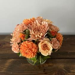 Short arrangement of orange and peach blooms in a clear glass bowl vase