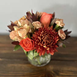 Short round arrangement of orange rose, rust chrysanthemum, and peach carnations in a glass vase