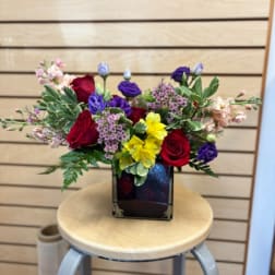 Mixed bouquet of red, purple, pink, and yellow flowers in a square vase