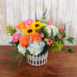 Low vase arrangement with orange roses, yellow sunflowers, pink gerbera daisies, tulips, and blue hydrangeas.