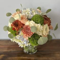 Round arrangement of peach roses and mums with white and blue hydrangeas in a clear glass vase