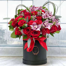 Red roses and pink orchids arranged in a black hatbox with a red ribbon