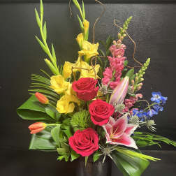 Mixed bouquet with red roses, yellow lilies, and pink blooms in a dark vase