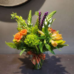 Orange roses and mixed blooms arranged in a glass vase with a plaid ribbon