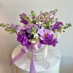 Purple and white floral arrangement in a white wicker basket with a lavender ribbon