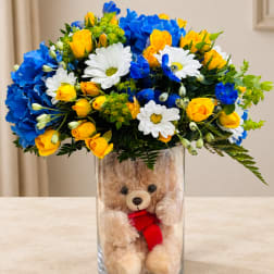 Bouquet of yellow roses, blue carnations, and white daisies in a glass vase with a teddy bear