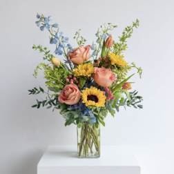 Tall mixed bouquet with yellow sunflowers, peach roses, blue blooms and tulips in a clear glass vase.