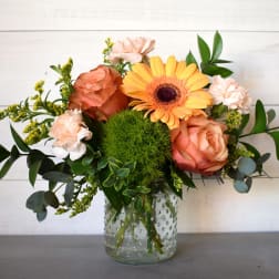 Peach roses, pale carnations, a yellow gerbera daisy, and a green pompom bloom in a clear glass jar.