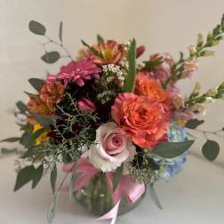 Mixed bouquet of pink, orange, and blue flowers in a glass vase with a pink ribbon