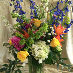 Tall mixed bouquet in a clear glass vase with blue, yellow, orange, and white flowers