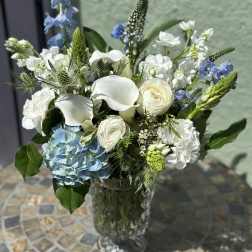 Arrangement of white calla lilies, roses, and blue hydrangea in a tall clear glass vase