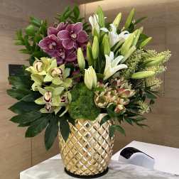 Tall arrangement of orchids, lilies and protea in a textured gold vase on a marble counter