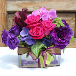 Purple and pink floral arrangement in a clear square vase with ribbon