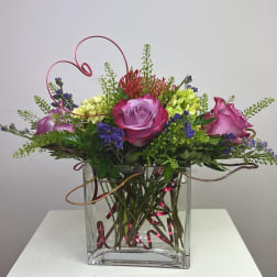 Pink and purple roses arranged in a clear square vase with curly wire accents