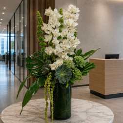 Tall white orchid arrangement in a dark glass vase with green tropical accents