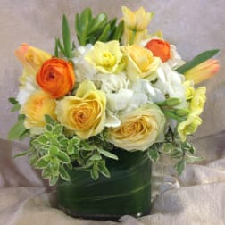 Compact arrangement of yellow roses, white hydrangeas, and orange blooms in a leaf-wrapped glass vase.