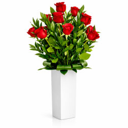 Red roses arranged in a tall white vase with green foliage