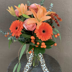 Orange lilies and gerbera daisies in a glass vase with a patterned ribbon