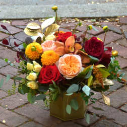 Autumn-tone arrangement of red roses, peach blooms and yellow flowers in a gold cube vase