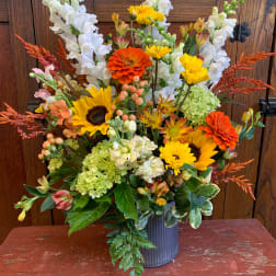 Mixed bouquet with sunflowers, orange zinnias, white snapdragons, and green hydrangeas in a metal vase