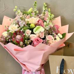 Large pink wrapped bouquet with pastel roses, protea, and mixed flowers beside a kraft shopping bag