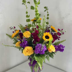 Mixed bouquet of yellow roses, sunflowers, and purple flowers in a glass vase
