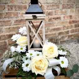 White roses and daisies arranged around a wooden lantern with a candle.