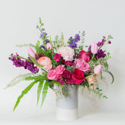 Asymmetrical pink and purple flower arrangement with tulips and roses in a tall white and gray cylinder vase