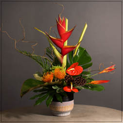 Tall tropical arrangement with red heliconia, orange blooms, and large leaves in a ceramic vase.