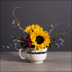 Compact arrangement of sunflowers, purple thistles, and red blooms in a white skyline mug