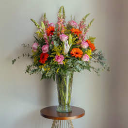 Tall arrangement of pink roses, orange gerbera daisies, lilies and snapdragons in a clear glass vase
