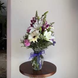 Tall mixed bouquet of white lilies and daisies with purple and pink flowers in a clear vase with a purple bow.