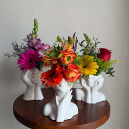 Three colorful flower arrangements in white face-shaped vases on a round table