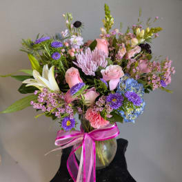 Mixed bouquet of pink, purple, and blue flowers in a glass vase with a pink ribbon