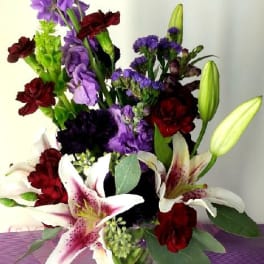 Mixed bouquet with lilies, purple blooms, and red carnations in a glass vase