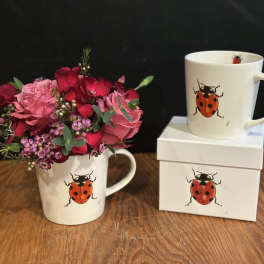 Pink and red flower bouquet in a ladybug mug beside matching gift boxes