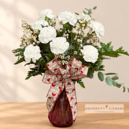 White carnations and filler flowers in a burgundy glass vase with a heart ribbon