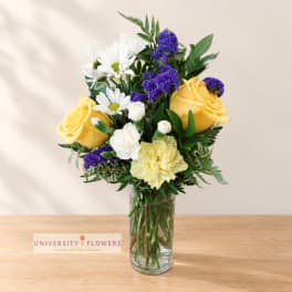 Yellow roses, white daisies, and purple filler flowers in a clear glass vase