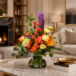 Mixed bouquet of orange, pink, yellow, and purple flowers in a glass vase