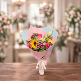 Mixed bouquet with sunflowers, roses, lilies, and purple daisies wrapped in pink and blue paper